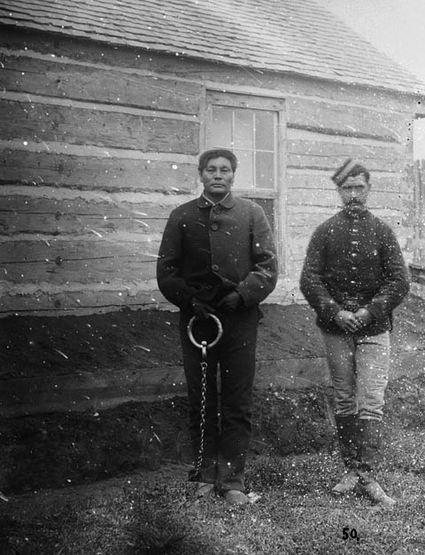 Black and white photograph of Swift Runner, shackled, next to another man.