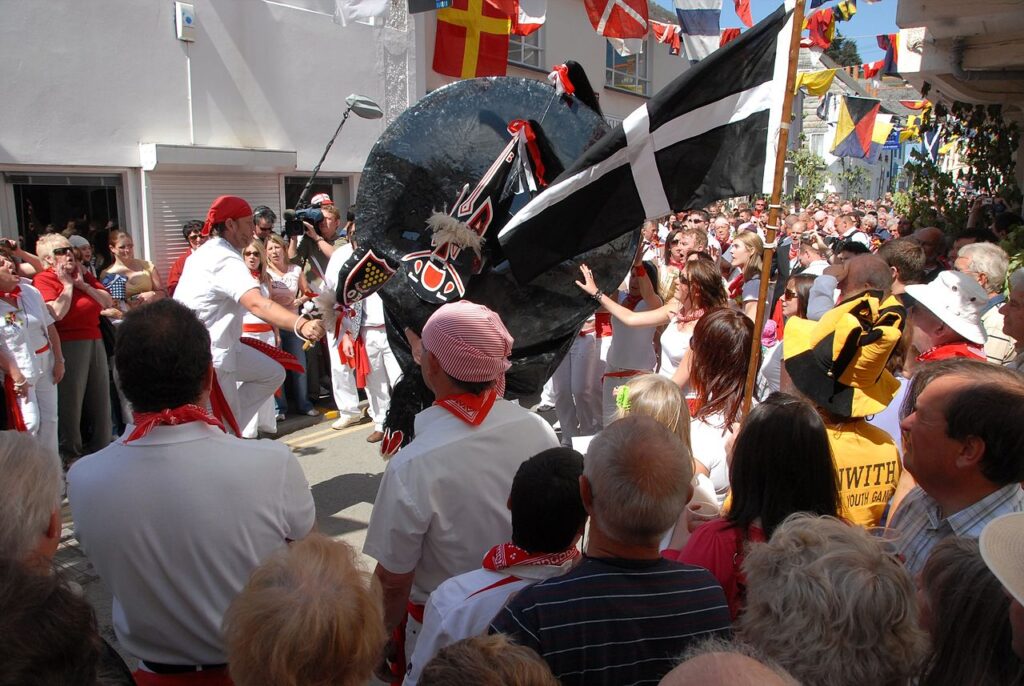 Padstow's old 'Obby 'Oss parades with the teaser and his club to the left. The tail of the 'Oss is at the top with the head pointed down. Teaser and 'Oss dance as they travel the streets amongst a crowd singing in praise of summer. Cornwall's national flag, St Piran's white cross on a black background, unfurls in the foreground.By Bryan Ledgard, CC BY 2.0, https://commons.wikimedia.org/w/index.php?curid=58269367