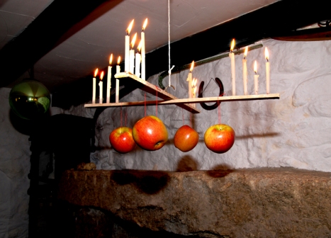 Hanging wooden cross, supporting lit candles with apples hanging from it on string.